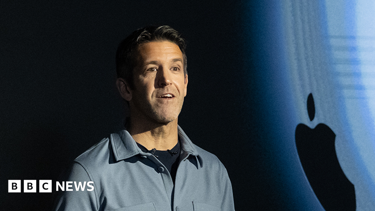 John Ternus standing on stage in front of an Apple logo. He is wearing a grey shirt.
