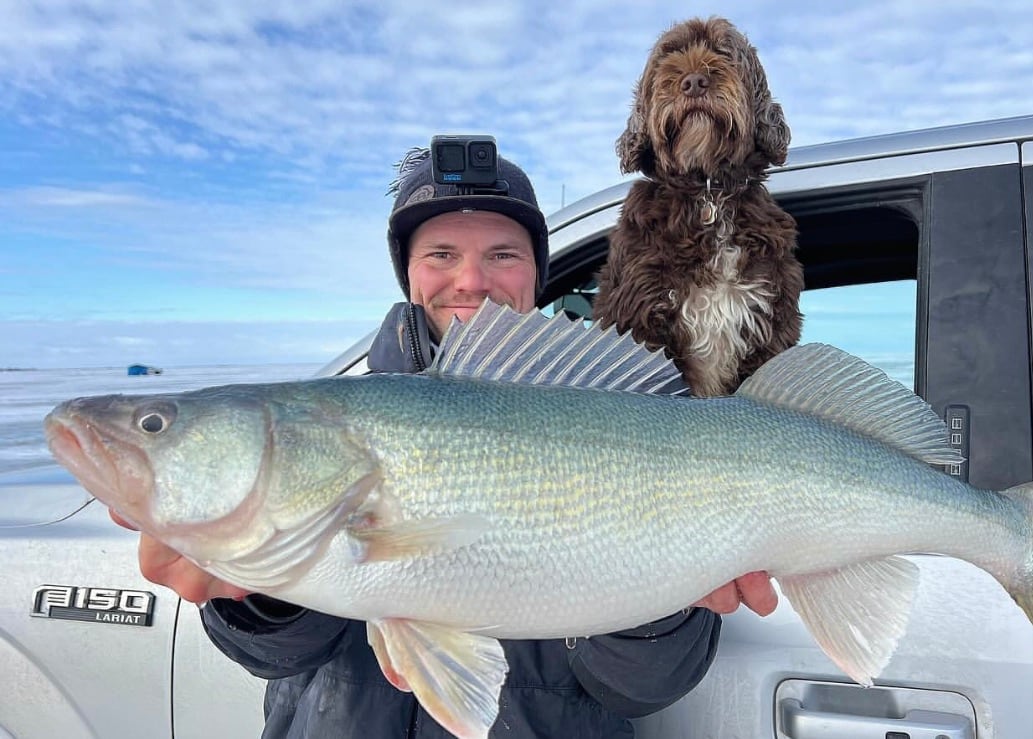 A man holding a fish in front a dog and a truck.