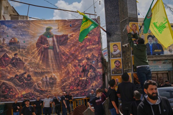 Hezbollah supporters hang portraits of fighters killed in Israeli airstrikes beside a large billboard depicting the war in the Middle East following U.S.-Israeli strikes on Iran on Feb. 28, ahead of a funeral procession in the southern village of Kfar Sir, Lebanon, Tuesday, April 21, 2026. (AP Photo/Hassan Ammar)