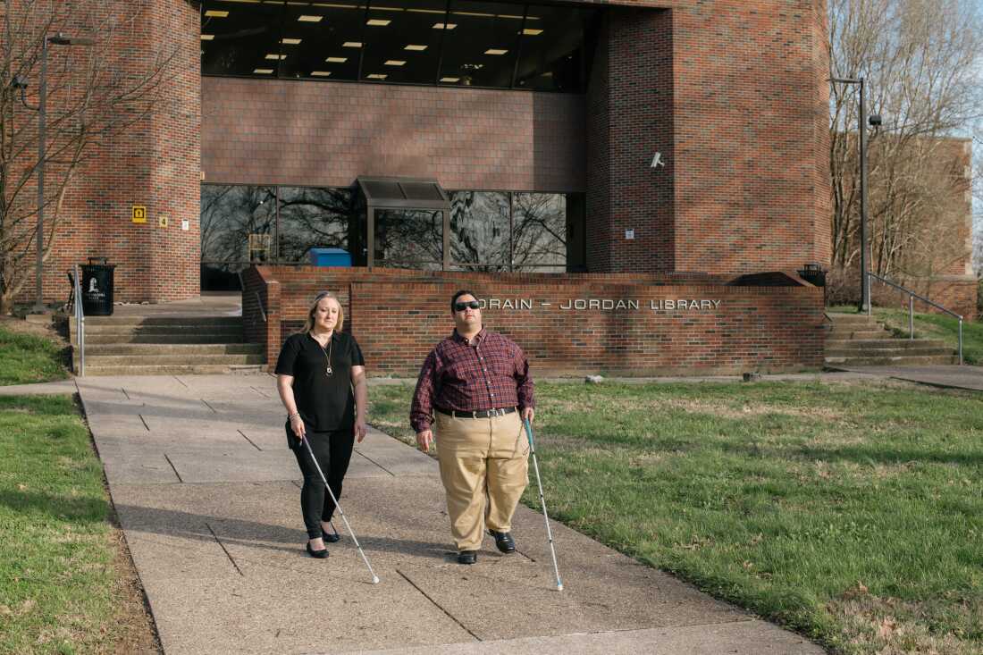 Two people who are blind --- a white, blonde woman with shoulder-length hair and a white man short, dark hair and sunglasses --- are walking on the campus of West Virginia State University while holding canes. 