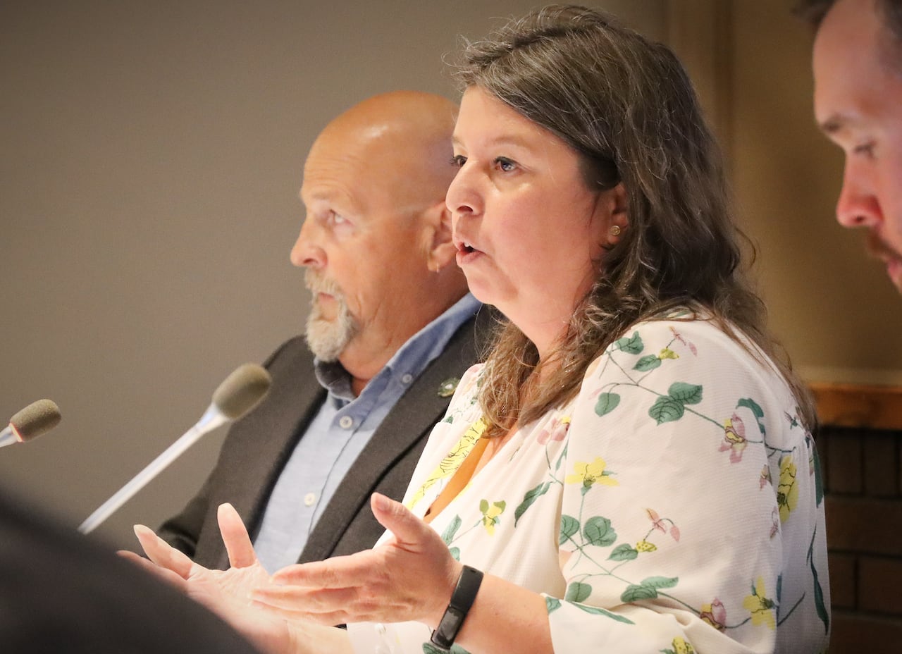 A woman in a white top with flower prints gestures with her hands as she speaks into a microphone.