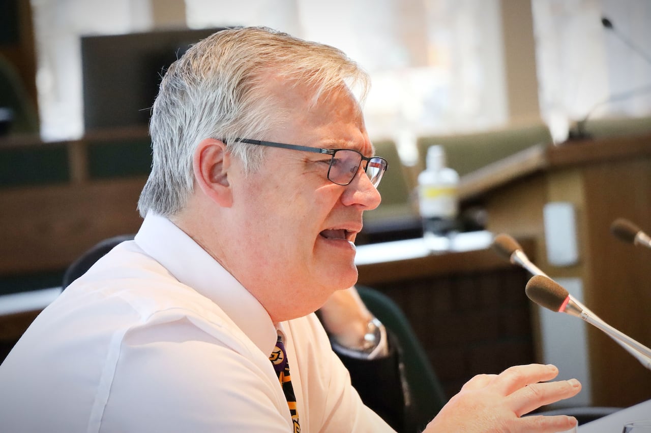 A man with grey hair, glasses and a white shirt gestures as he speaks into a microphone.