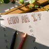 A photograph of a piece of paper in a journal that has "Who Am I?" written in block letters at the top of the page. Below it are two columns of numbers from 1-20. The notebook has an open black pen lying on top of the page, and lays on a wooden slatted surface with a vine of Swedish ivy framing the book overhead. 