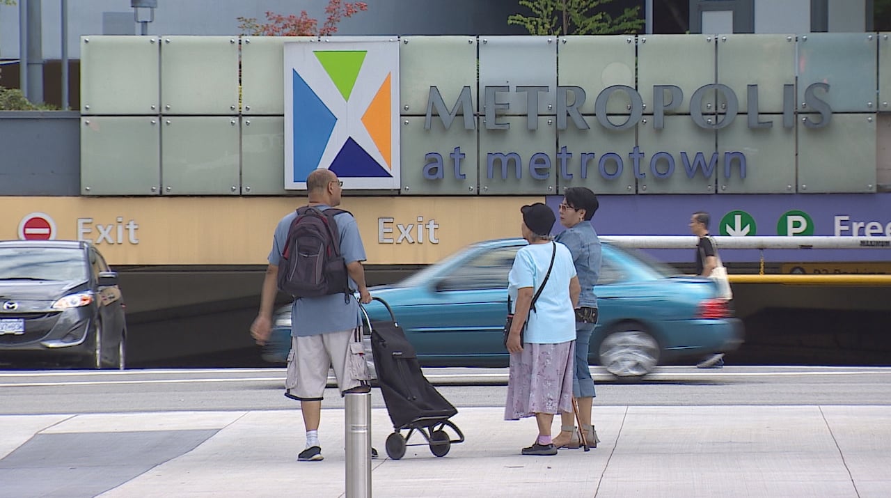 People walk by a sign that reads 'Metropolis at Metrotown' with cars zooming by.