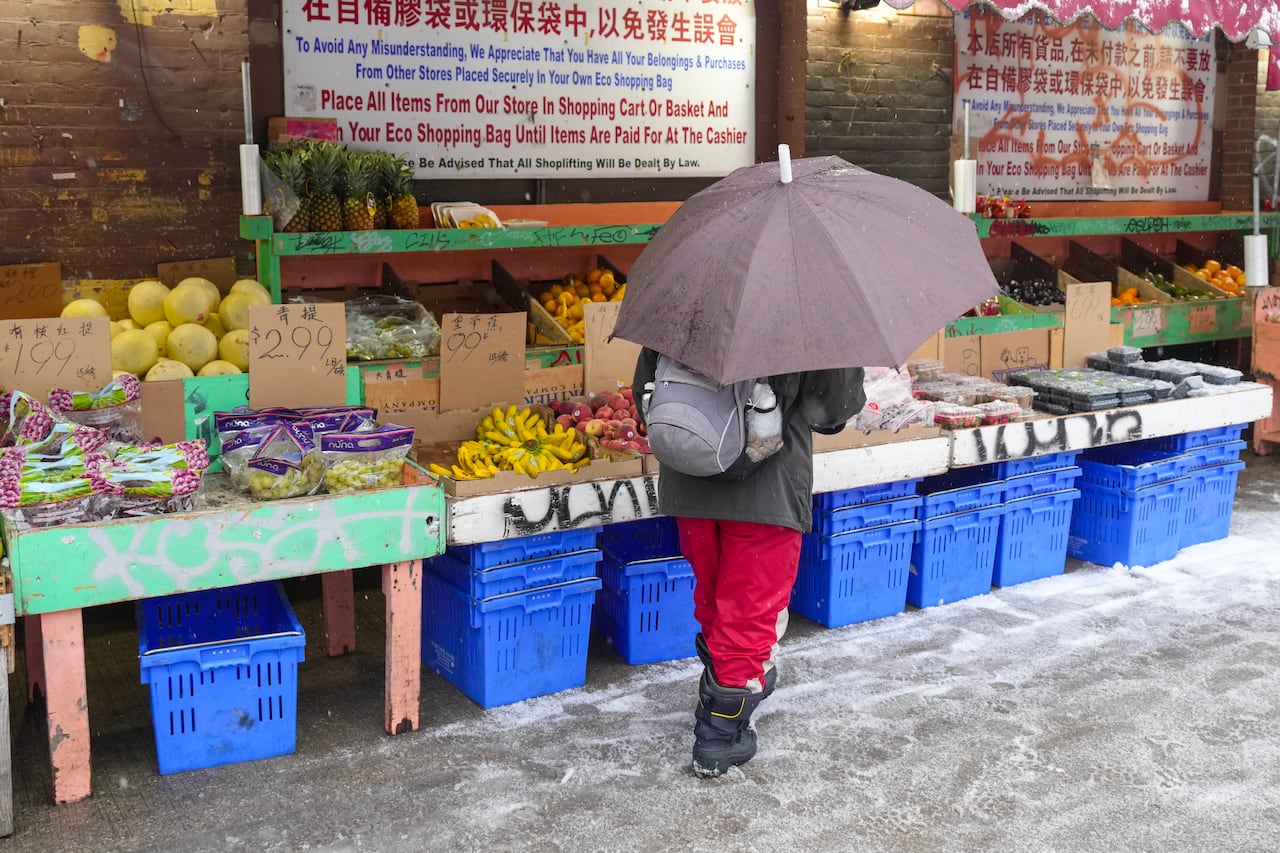 A man stands in front of an outdoor fruit stand on a city sidewalk