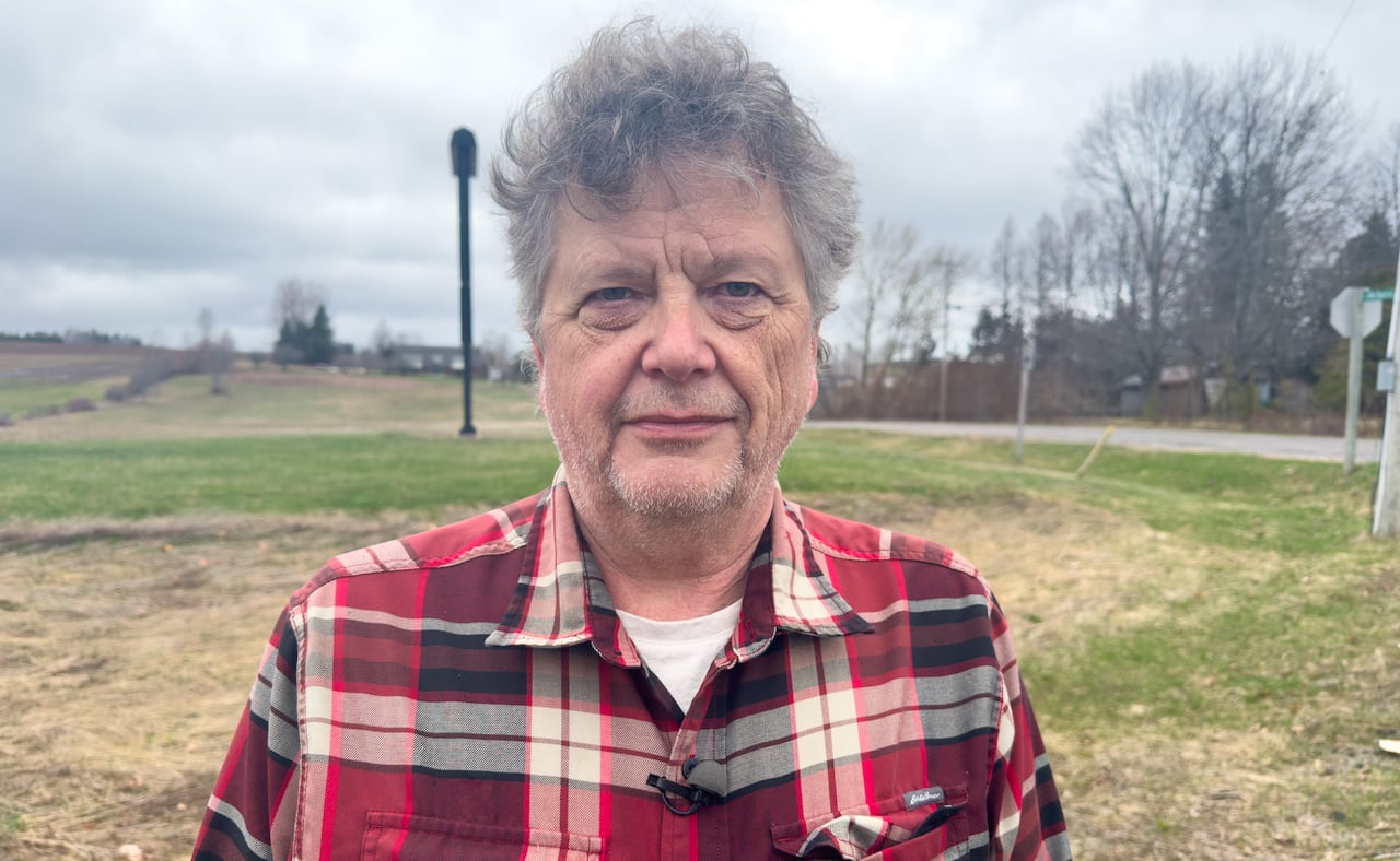 A man with grey hair, wearing a red plaid shirt, standing in a dry field