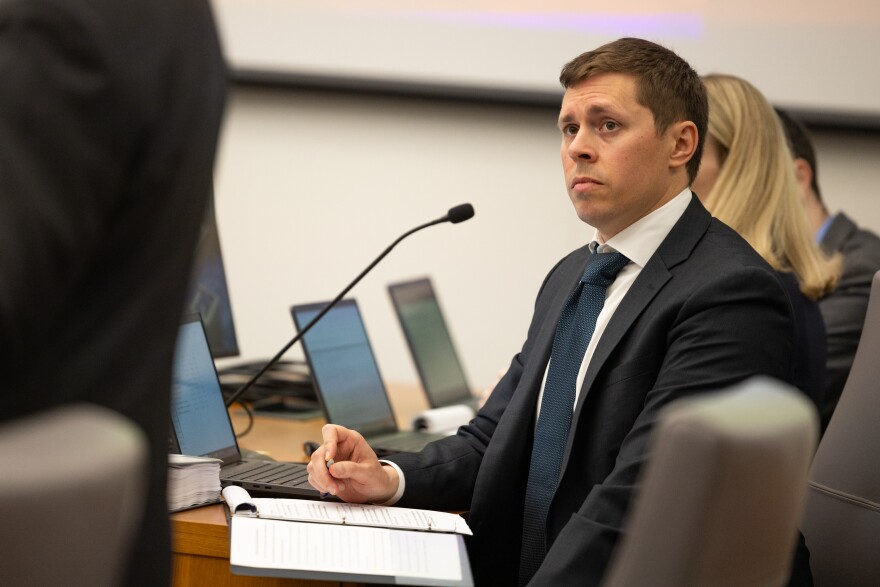 A man in a dark suit sits at a table with a laptop open in front of him while watching another person speak at a courtroom podium.