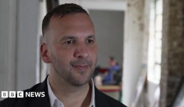 Zack Polanski being interviewed for TV. He is wearing a dark jacket and a white open-necked shirt. There is an blurred background image with a window to one side.