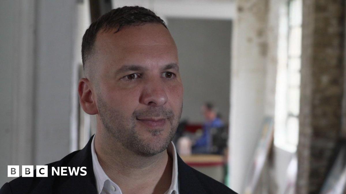 Zack Polanski being interviewed for TV. He is wearing a dark jacket and a white open-necked shirt. There is an blurred background image with a window to one side.