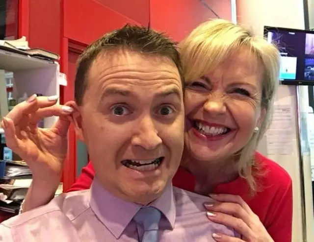 Carol playfully pulling Matt’s ear, both smiling with humorous expressions in a newsroom filled with bright lights, desks and equipment