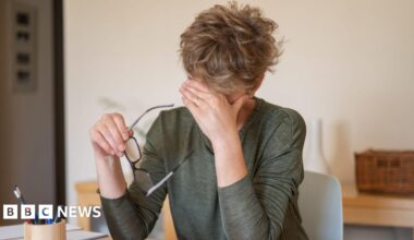 A woman sat at a desk rubbing her forehead holding a pair of glasses.