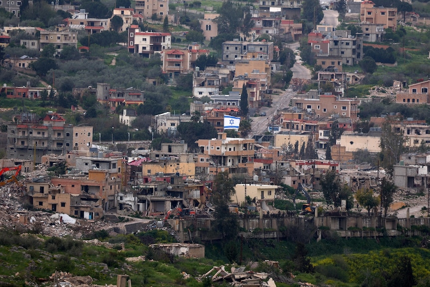 a view of a hillside village with many houses demolished with heavy machinery operating and an Israeli flag in the centre