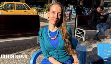 Shelly Kittleson, who has long dark blonde hair and wears a blue dress with a turquiose cardigan, sits on a plastic chair in the sun in a Baghdad street