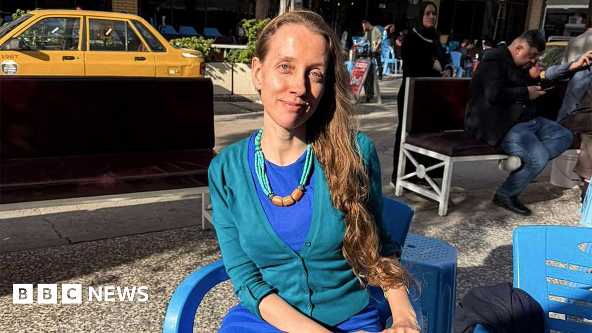 Shelly Kittleson, who has long dark blonde hair and wears a blue dress with a turquiose cardigan, sits on a plastic chair in the sun in a Baghdad street