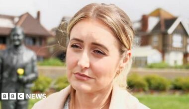 A blonde woman standing, looking past the camera. There is a metal statue over her right shoulder holding a yellow flower.
