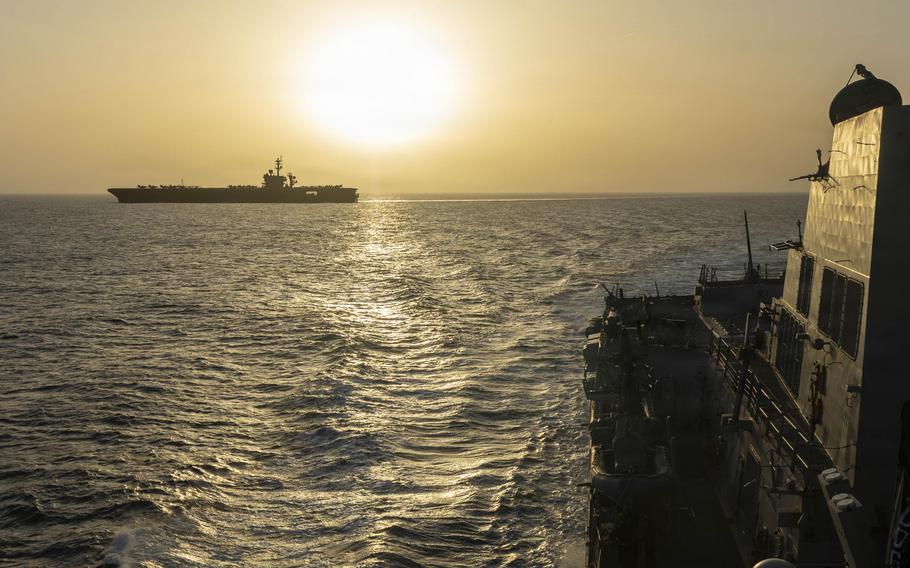 An aircraft carrier silhouetted against the horizon, viewed from the deck of another ship at sea.