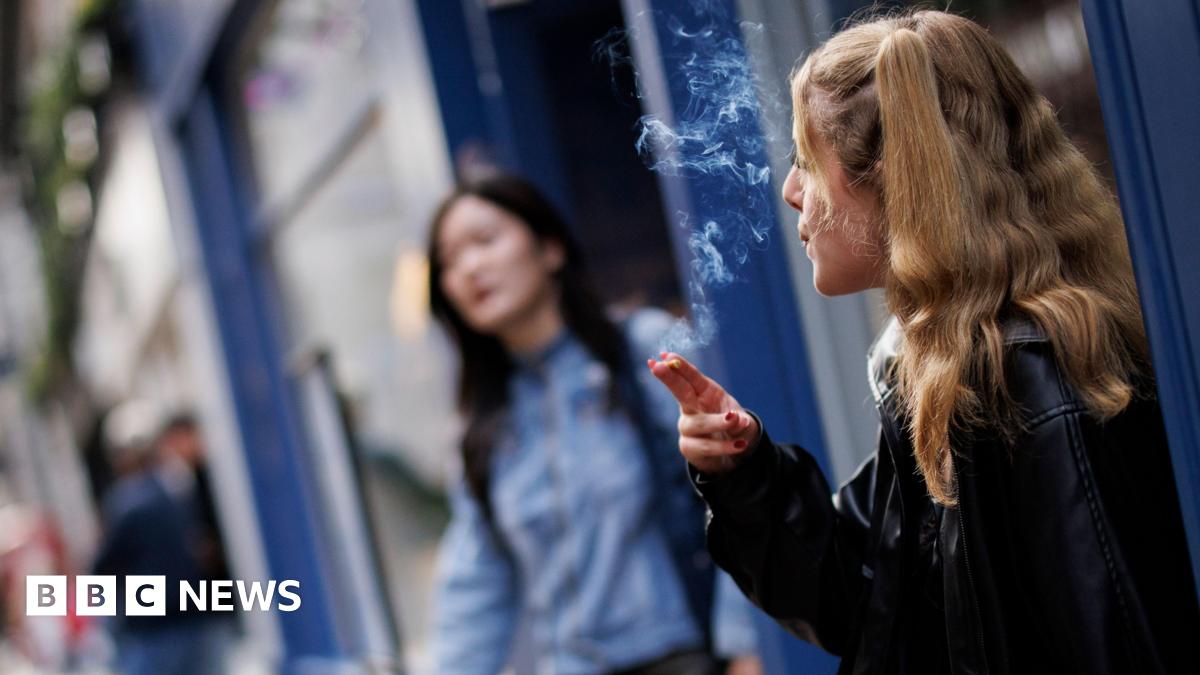 A young woman smokes outside a shop. She has long blonde hair and wears a leather jacket