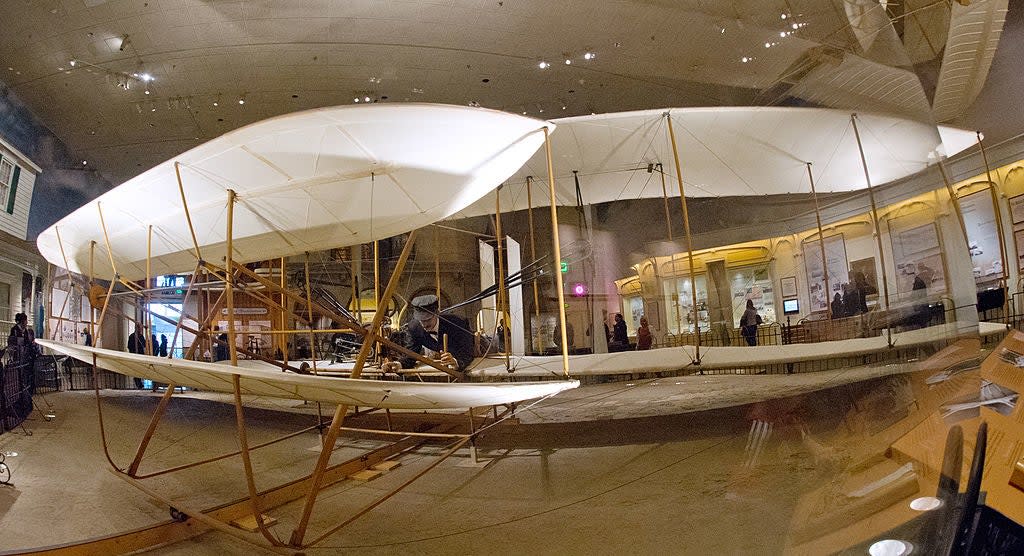The image shows the Wright brothers' early airplane on display in a museum exhibit, capturing a pioneering moment in aviation history