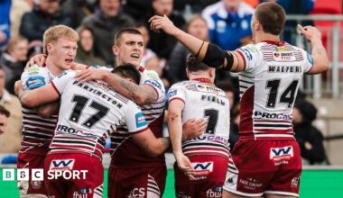 Wakefield's Tom Johnstone diving towards the line to score a try one-handed for his side under pressure from two Wigan defenders in the corner.