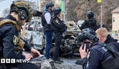 Six Ukrainian experts wearing helmets, and some of them wearing dark blue uniforms, work at the site of a drone attack in the Ukrainian city of Kharkiv. Behind them stands a wrecked car.