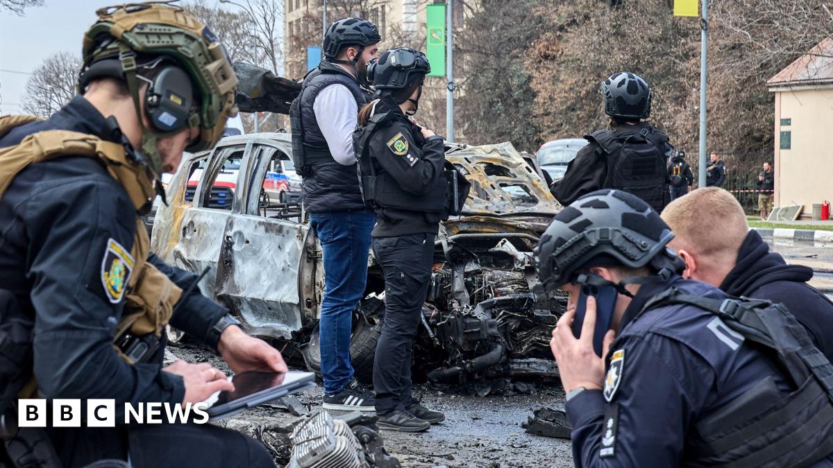 Six Ukrainian experts wearing helmets, and some of them wearing dark blue uniforms, work at the site of a drone attack in the Ukrainian city of Kharkiv. Behind them stands a wrecked car.