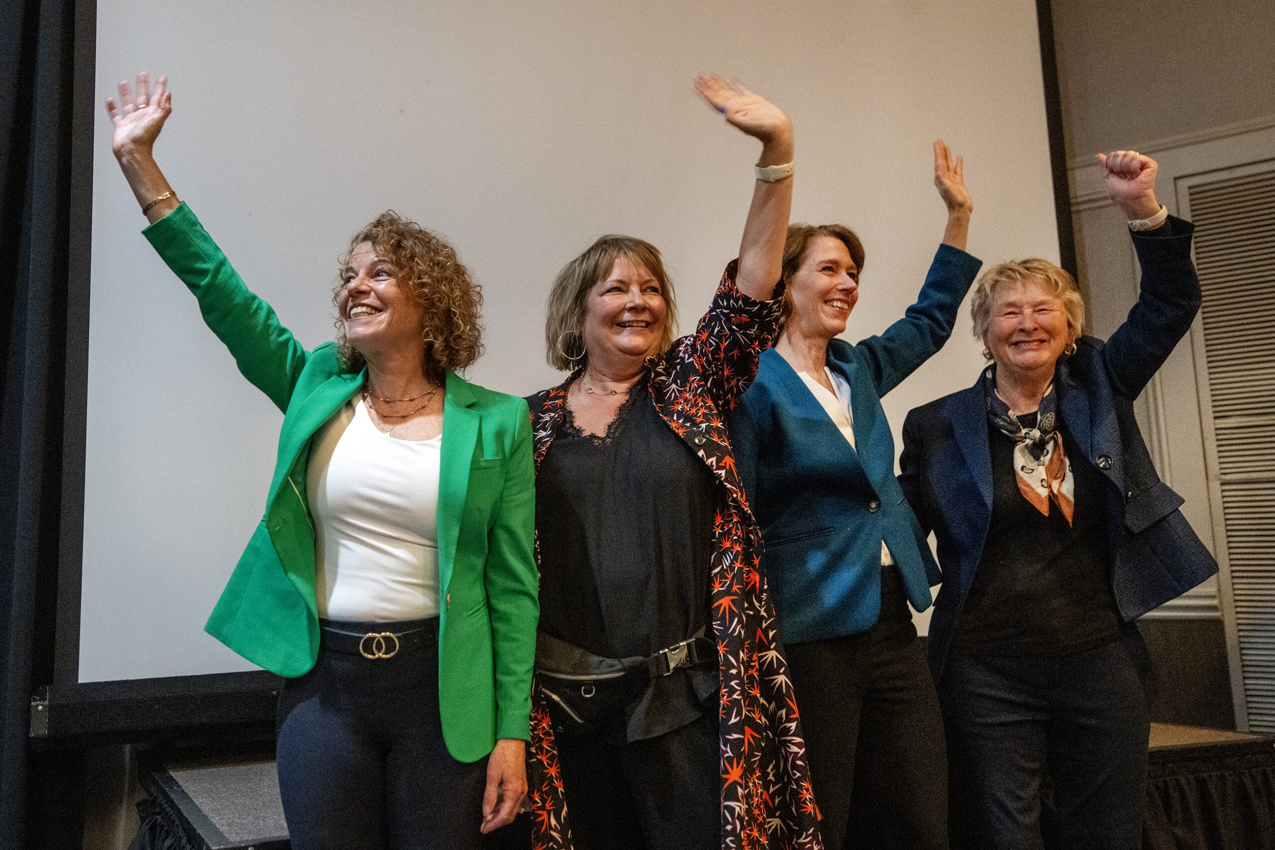 Four women stand side by side, smiling and waving in front of a blank projection screen in a conference room.
