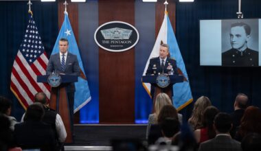 Two men, one in a business suit and the other in a military dress uniform, stand at lecterns indoors, with flags and a screen showing a person in the background.
