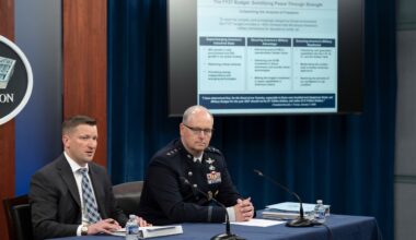 Two men, one in a suit and another in a military dress uniform, sit at a table. Behind them is a display screen showing a slide.