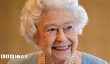 The late Queen. She is smiling and is wearing a duck-egg blue dress, a pearl necklace and pearl earrings.
