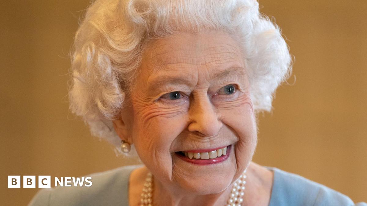 The late Queen. She is smiling and is wearing a duck-egg blue dress, a pearl necklace and pearl earrings.
