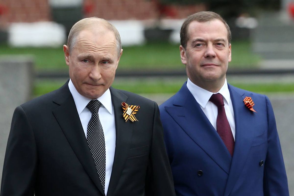 Two men in formal attire, one wearing a black suit and the other in a blue suit, standing side by side with a background that includes a grassy area and a concrete structure.