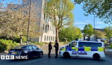 Police tape closes off the road. Two officers can be seen stood next to a police vehicle. The area is leafy with a pale building in the background.