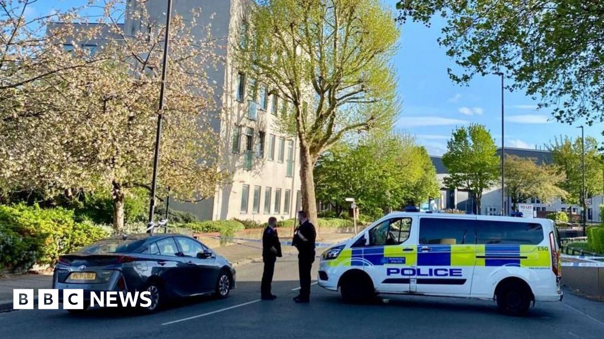 Police tape closes off the road. Two officers can be seen stood next to a police vehicle. The area is leafy with a pale building in the background.
