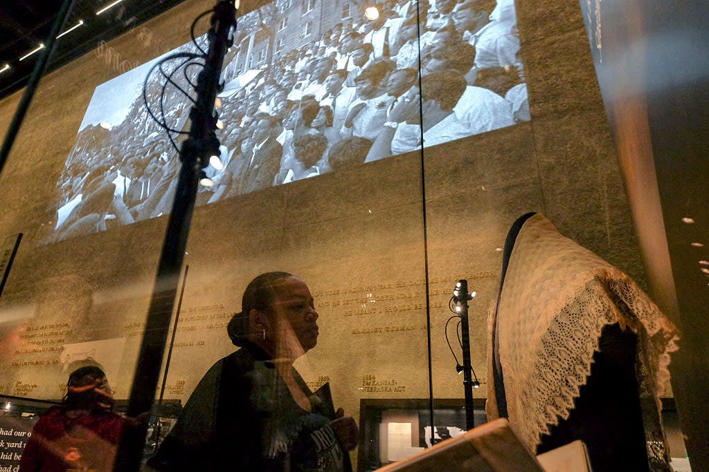 Museum exhibit with historical footage projected on a wall. A visitor observes a display case with vintage clothing
