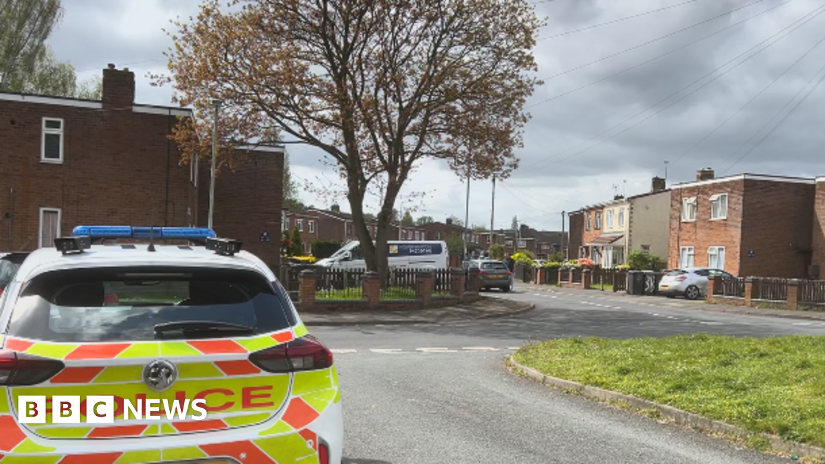 A police car is parked outside properties in Willis Pearson Avenue. A grass verge can be seen on the right with apartments/small blocks of flats visible off to the right and left of the image. A large stands outside one block.