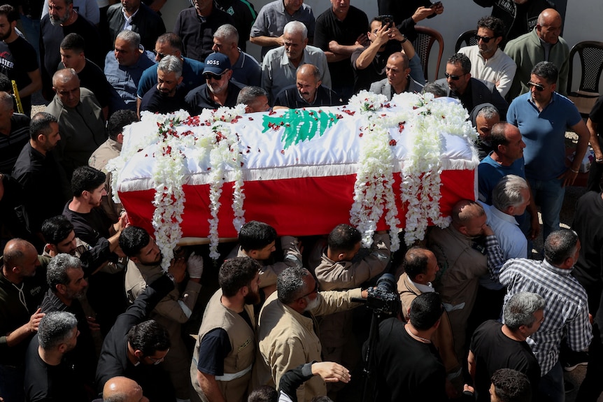 Image of a coffin covered in a shroud and flowers being carried through a crowd of people. 