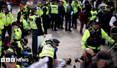 A large group of police officers form a circle around a man in a grey sweatshirt lying on the floor.