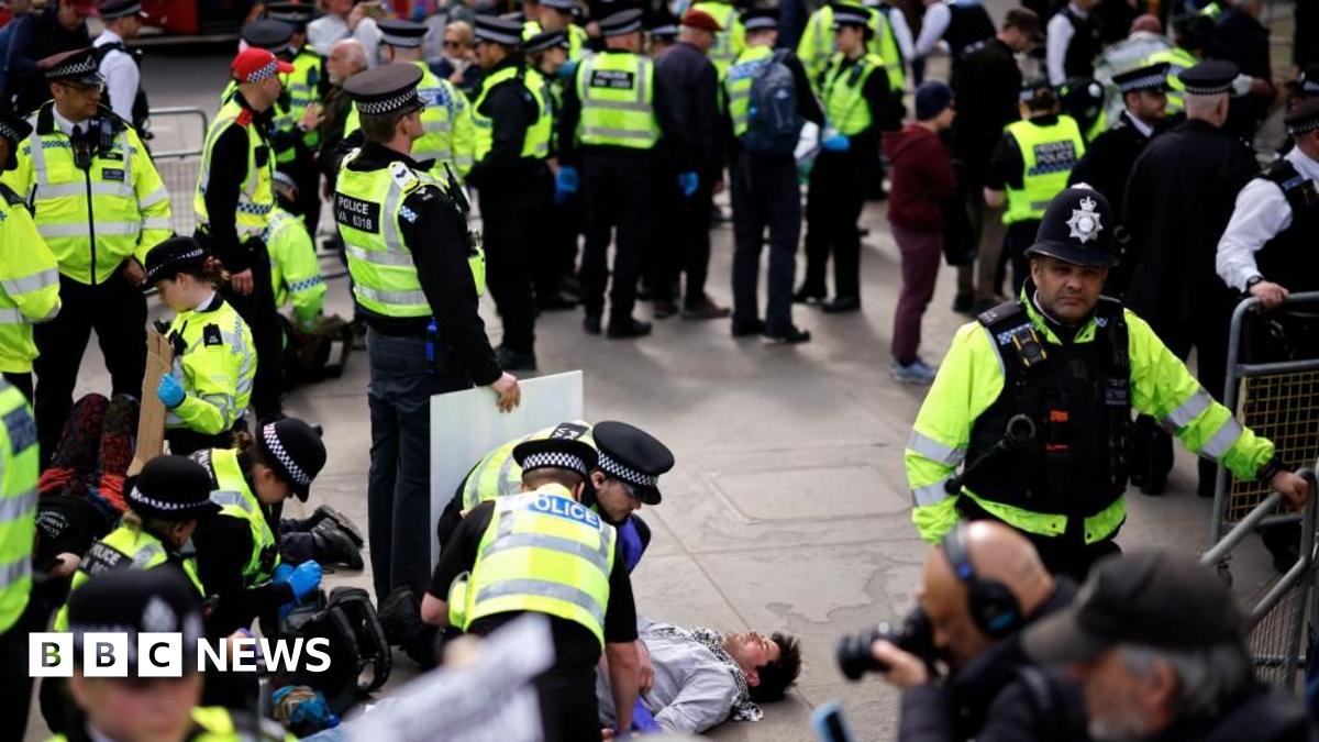 A large group of police officers form a circle around a man in a grey sweatshirt lying on the floor.