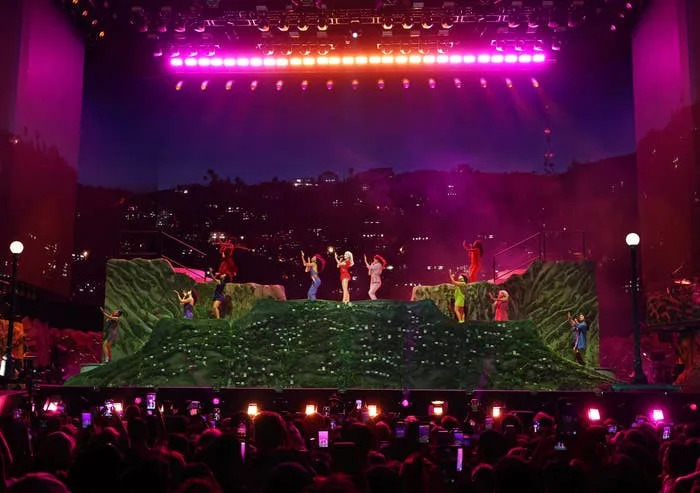 Performers on stage with a hillside set and cityscape backdrop, lit by bright stage lights. Crowd watching and taking photos