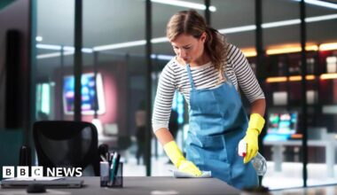 A woman working as a cleaner in an office building. She is wearing yellow rubber gloves, a striped top and a blue apron.