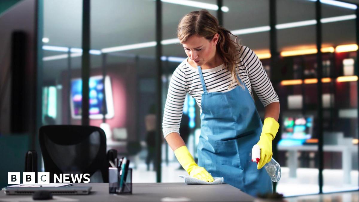 A woman working as a cleaner in an office building. She is wearing yellow rubber gloves, a striped top and a blue apron.