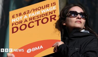 Seven people, five men and one woman stand in a lifeboat shed looking at the camera. A small lifeboat is in the background