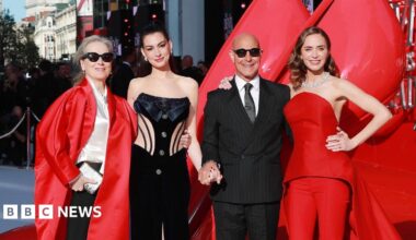 Meryl Streep, Anne Hathaway, Stanley Tucci and Emily Blunt pose on the red carpet in London. Streep is wearing sunglasses, a long, red, satin jacket, a cream blouse and black trousers. Hathaway has her hair up and is wearing a black embellished corset. Tucci is wearing a pinstriped black suit, white shirt and tie, along with sunglasses. Blunt has a red two-piece on an a jewelled necklace.
