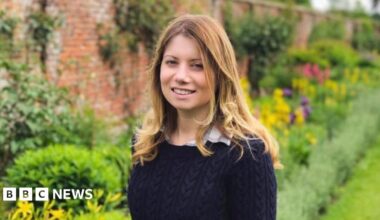 Kevin Miller is wearing a grey or navy blue suit while walking on a pavement and carrying a dark holdall in his left hand.