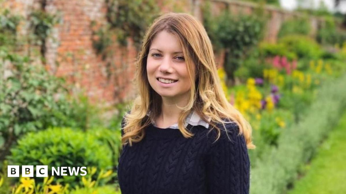 Kevin Miller is wearing a grey or navy blue suit while walking on a pavement and carrying a dark holdall in his left hand.