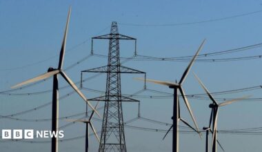 Six wind turbines in front of electricity pylons, with a light blue sky in the background.