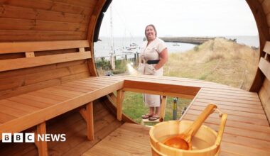 The image shows the inside of a wooden style sauna, Kathryn Donavan, the owner of the sauna is stood just outside. She's wearing a beige dress with a black belt and a pair of black flip flops. She has short blonde hair.