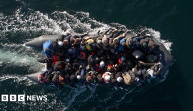 Dozens of people are crammed onto a small black dinghy, which leaves white trails of foam behind it in the sea