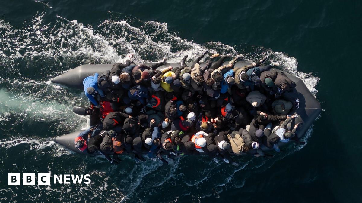 Dozens of people are crammed onto a small black dinghy, which leaves white trails of foam behind it in the sea