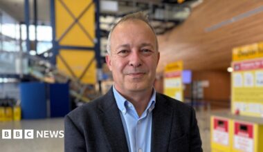 Nico Bezuidenhout, a man with short grey hair, wearing a light blue shirt and a dark blazer. He is smiling directly at the camera, while standing in the departures hall at Guernsey Airport.
